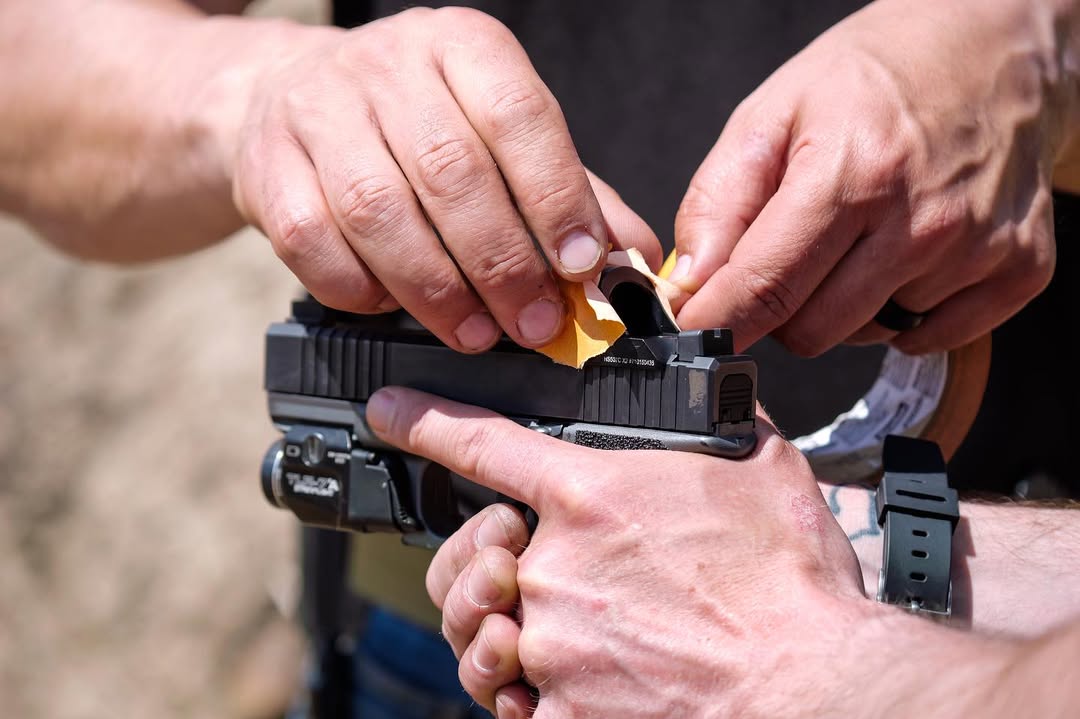 Man taping the front of a red dot sight on a handgun for training purposes.